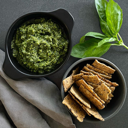 Bowl of green pesto with crackers and basil leaves on a dark surface