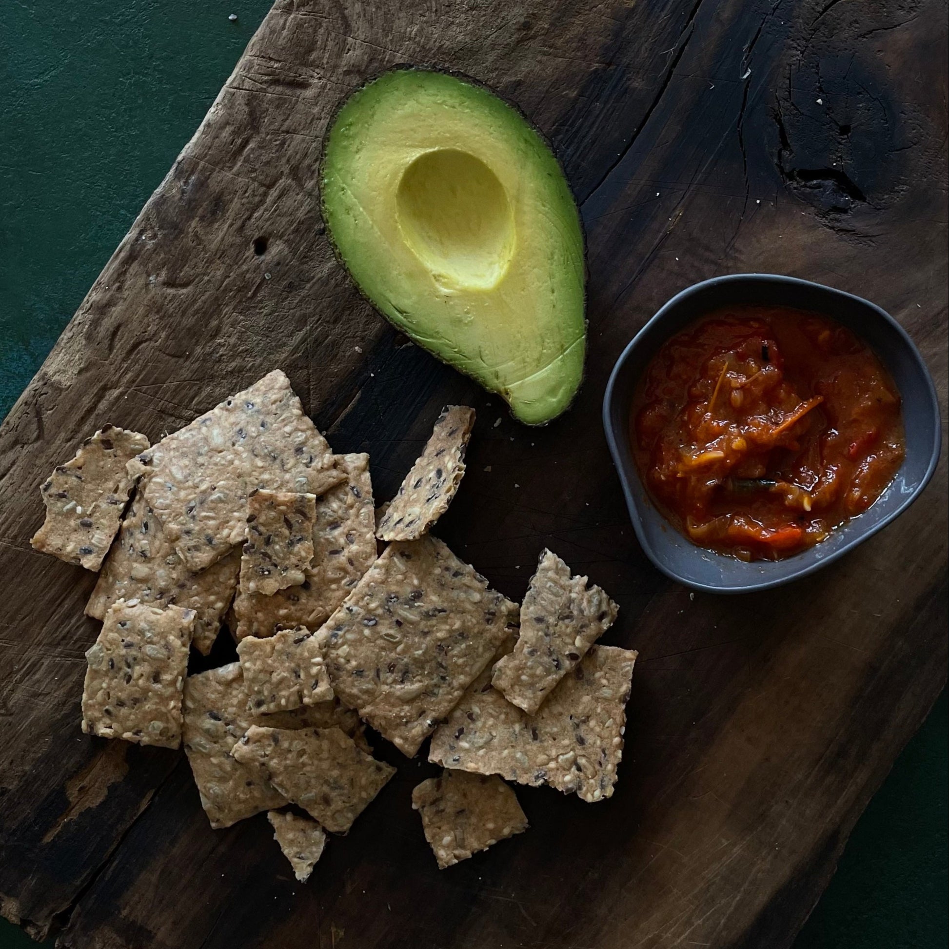 Avocado half, seed crackers, and tomato relish on a wooden board