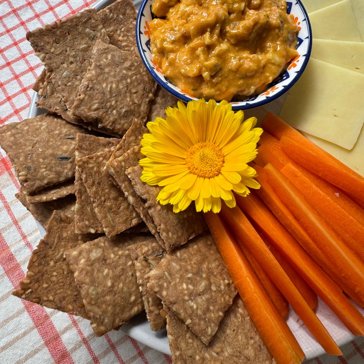 Plate of Smoked Paprika crackers with carrot sticks, sliced cheese, an orange dip and a yellow flower